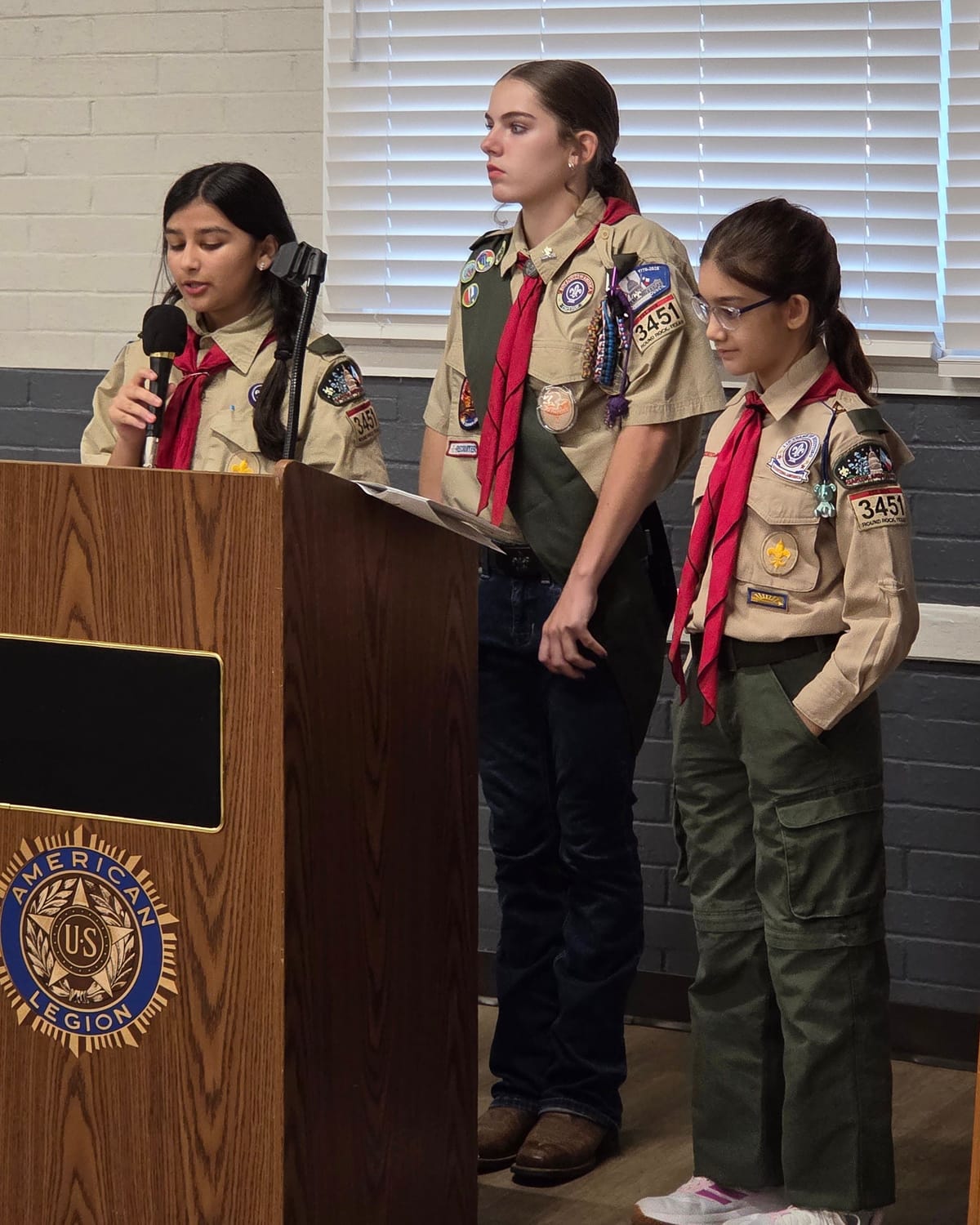 Viti speaking into a microphone as emcee, with two fellow scouts beside her
